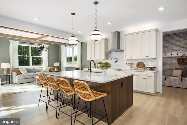 kitchen featuring wall chimney range hood, sink, white cabinetry, hanging light fixtures, and a kitchen island with sink