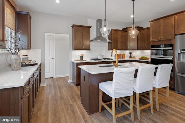 kitchen featuring stainless steel refrigerator with ice dispenser, decorative light fixtures, a center island with sink, light stone countertops, and wall chimney range hood