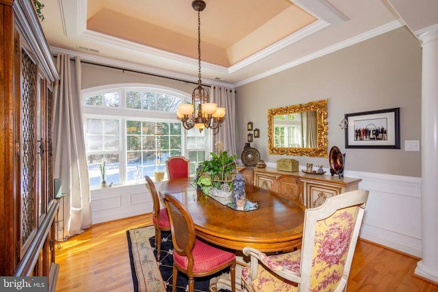 dining room featuring a wainscoted wall, ornate columns, a raised ceiling, light wood-type flooring, and a chandelier