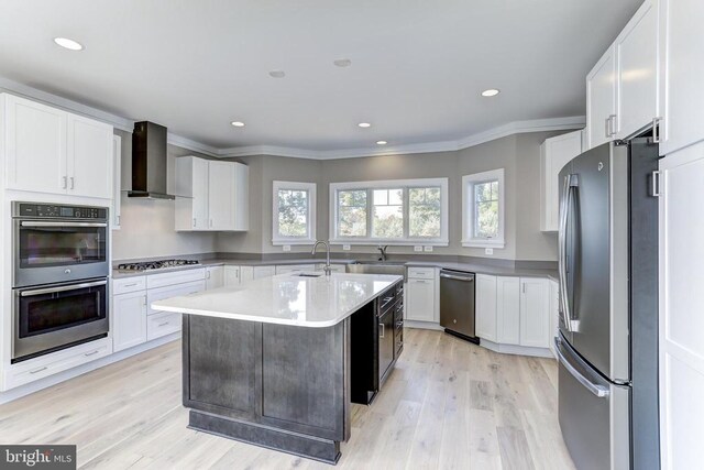 kitchen with stainless steel appliances, an island with sink, white cabinets, wall chimney exhaust hood, and light wood-type flooring