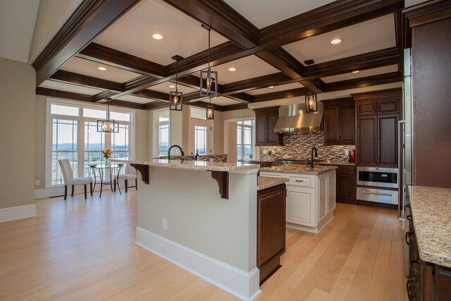 kitchen featuring pendant lighting, light hardwood / wood-style flooring, a kitchen breakfast bar, an island with sink, and wall chimney exhaust hood