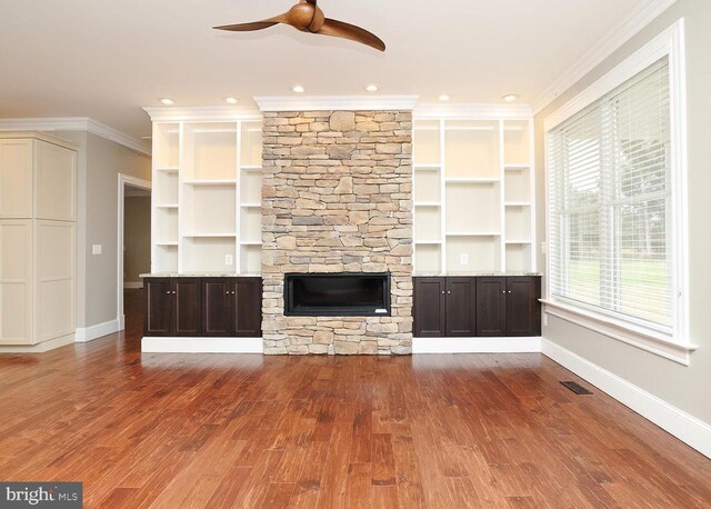 unfurnished living room with ceiling fan, ornamental molding, a stone fireplace, and wood-type flooring