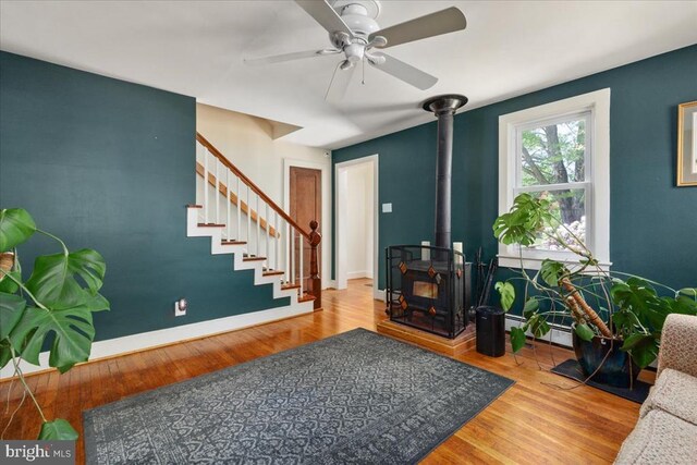 living area with a wood stove, stairs, wood finished floors, baseboards, and a ceiling fan