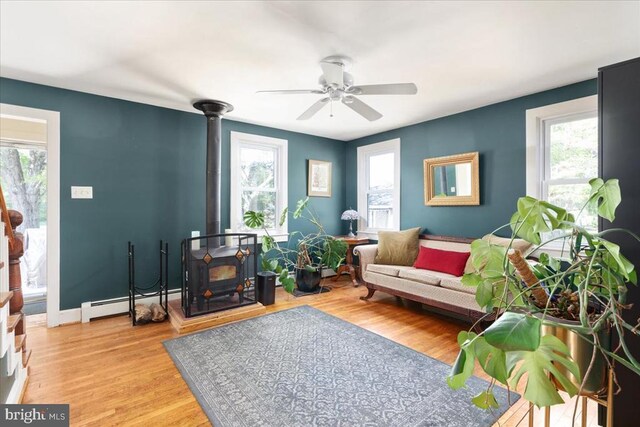 living area with ceiling fan, a healthy amount of sunlight, a wood stove, and a baseboard radiator