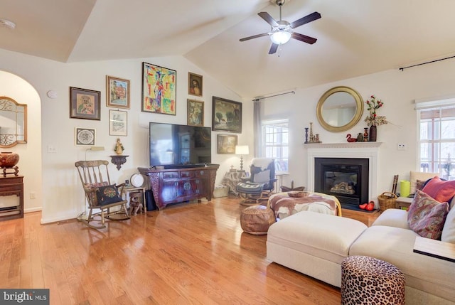 living room featuring light wood finished floors, lofted ceiling, arched walkways, a glass covered fireplace, and a ceiling fan