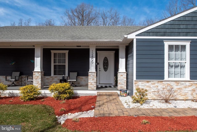 view of exterior entry with stone siding, covered porch, and a shingled roof