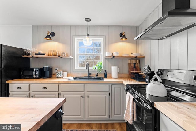 kitchen with wooden counters, a sink, black appliances, wood finished floors, and wall chimney exhaust hood