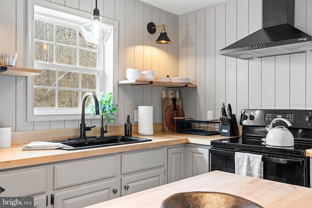 kitchen with butcher block counters, wall chimney range hood, black range with electric cooktop, open shelves, and a sink