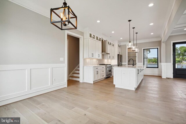 kitchen featuring wall chimney range hood, appliances with stainless steel finishes, an island with sink, white cabinets, and decorative light fixtures