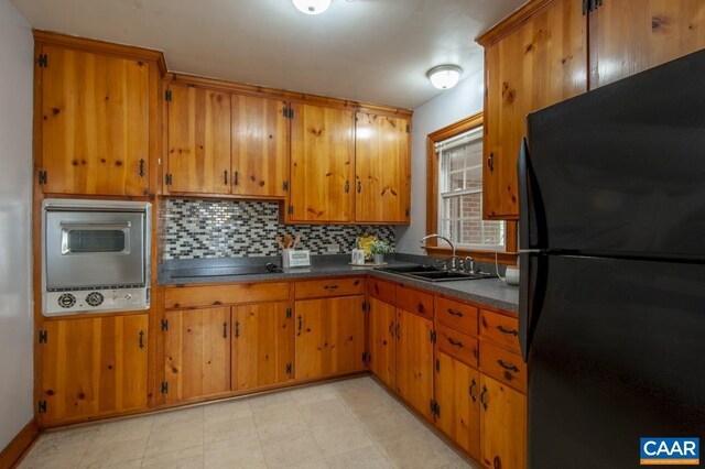 kitchen featuring a sink, backsplash, freestanding refrigerator, brown cabinets, and dark countertops