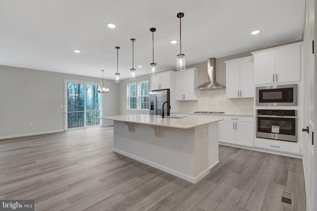 kitchen featuring a center island with sink, hanging light fixtures, stainless steel appliances, light countertops, and wall chimney range hood