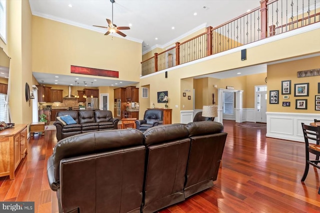 living room featuring a wainscoted wall, wood finished floors, a ceiling fan, a towering ceiling, and crown molding