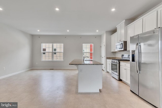 kitchen featuring a center island with sink, stainless steel appliances, white cabinets, a sink, and dark stone counters