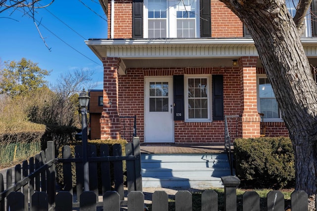 doorway to property with covered porch, brick siding, and fence