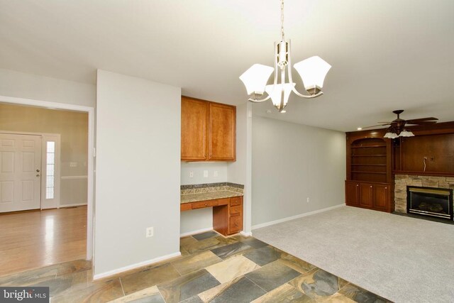 kitchen featuring a stone fireplace, built in desk, carpet flooring, pendant lighting, and ceiling fan with notable chandelier