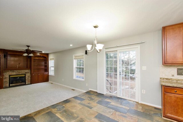 interior space featuring a stone fireplace, ceiling fan with notable chandelier, tasteful backsplash, hanging light fixtures, and carpet