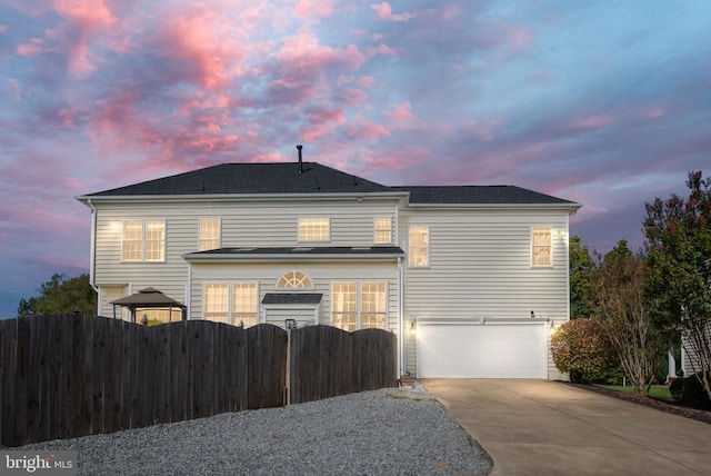 back of property at dusk featuring concrete driveway, an attached garage, fence, and a gate