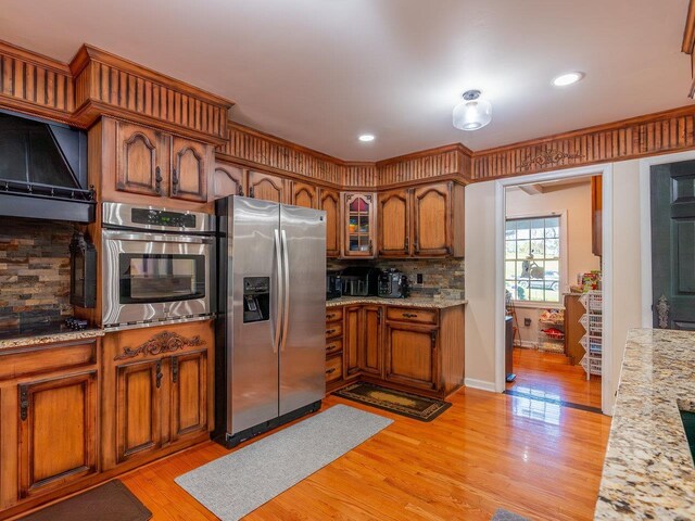 kitchen with light stone counters, tasteful backsplash, light wood-type flooring, appliances with stainless steel finishes, and range hood