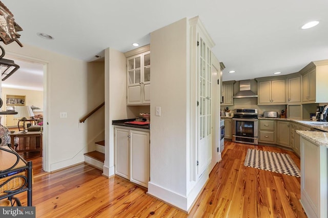 kitchen featuring wall chimney range hood, stainless steel electric range, light stone counters, light wood-style floors, and glass insert cabinets