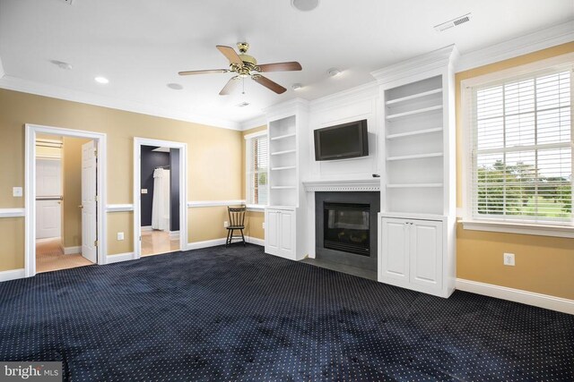 unfurnished living room featuring dark carpet, ornamental molding, built in shelves, and ceiling fan