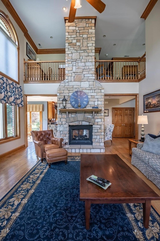 living room featuring a stone fireplace, wood-type flooring, ornamental molding, a towering ceiling, and ceiling fan