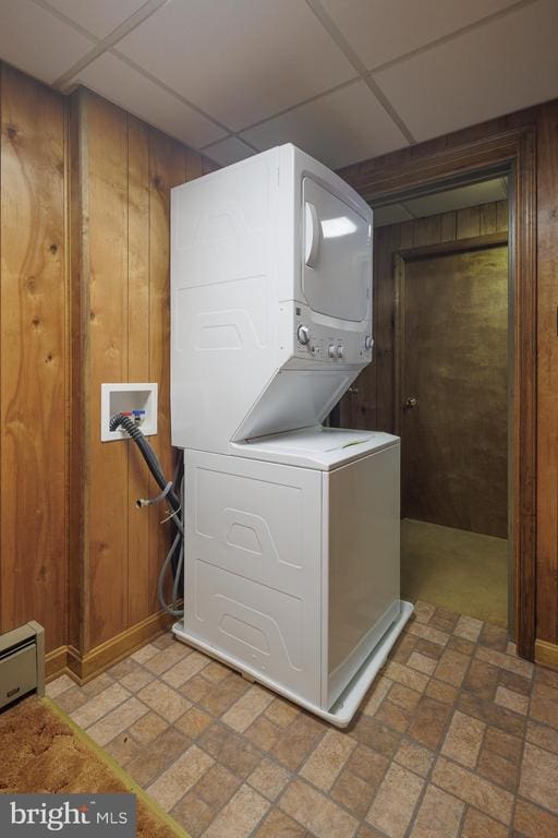 washroom featuring stacked washer / drying machine, wood walls, and a baseboard heating unit