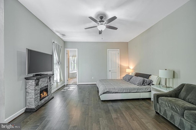 bedroom with a stone fireplace, a ceiling fan, dark wood-type flooring, and baseboards