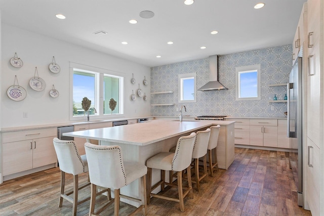 kitchen featuring a kitchen breakfast bar, a kitchen island with sink, dark hardwood / wood-style flooring, light brown cabinetry, and wall chimney range hood