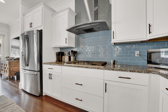 kitchen with wall chimney range hood, appliances with stainless steel finishes, dark stone countertops, white cabinets, and dark hardwood / wood-style flooring