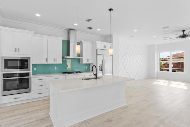 kitchen with ceiling fan, wall chimney range hood, tasteful backsplash, an island with sink, and built in appliances