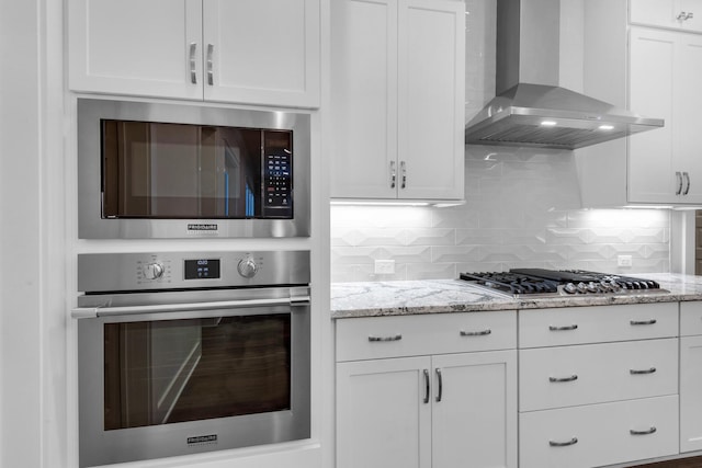 kitchen featuring white cabinets, backsplash, light stone countertops, stainless steel appliances, and wall chimney range hood