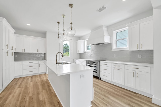 kitchen with custom exhaust hood, a center island with sink, sink, stainless steel range, and white cabinetry