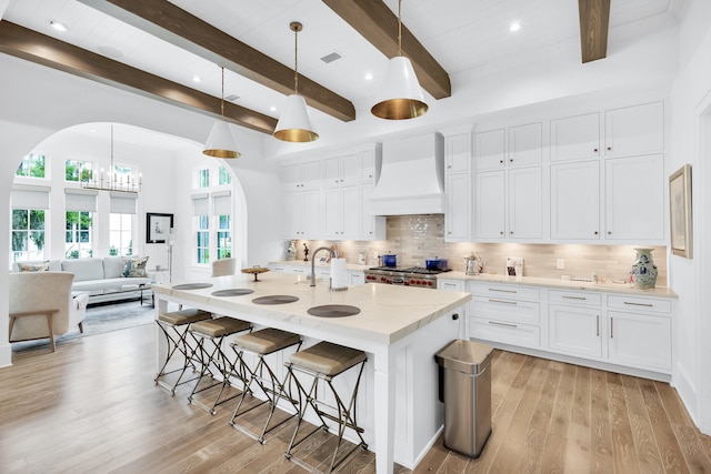 kitchen featuring light hardwood / wood-style floors, beamed ceiling, custom range hood, a kitchen island with sink, and white cabinets