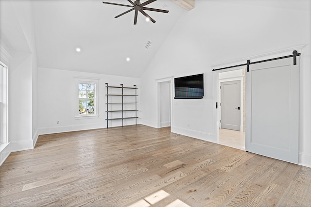 unfurnished living room with beam ceiling, light wood-type flooring, high vaulted ceiling, a barn door, and ceiling fan