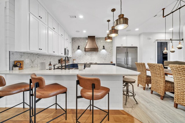 kitchen with tasteful backsplash, hanging light fixtures, light hardwood / wood-style flooring, premium range hood, and built in appliances