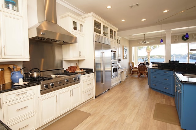 kitchen featuring built in appliances, hanging light fixtures, ceiling fan, wall chimney range hood, and blue cabinets