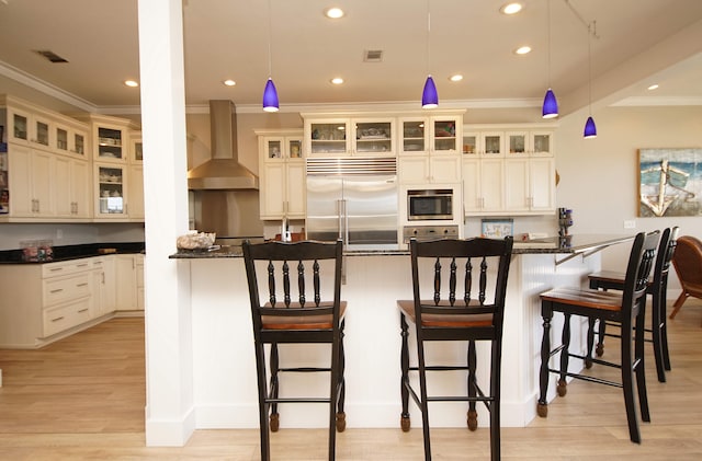 kitchen featuring built in appliances, wall chimney range hood, decorative light fixtures, and a breakfast bar area