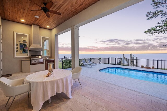 pool at dusk with a patio area, a water view, exterior kitchen, area for grilling, and ceiling fan