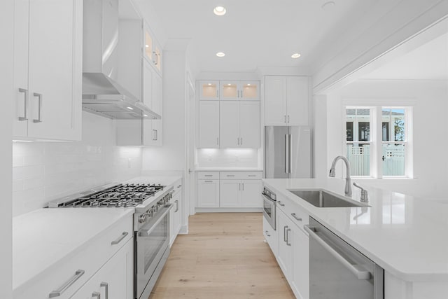 kitchen with appliances with stainless steel finishes, light countertops, light wood-type flooring, wall chimney range hood, and a sink