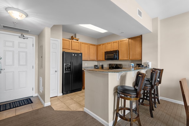 kitchen featuring kitchen peninsula, stone countertops, black appliances, a kitchen breakfast bar, and light tile floors