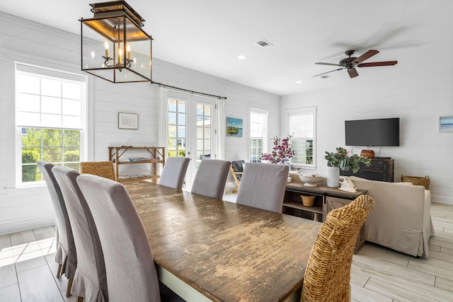 dining space with ceiling fan with notable chandelier, wood walls, french doors, and light wood-type flooring