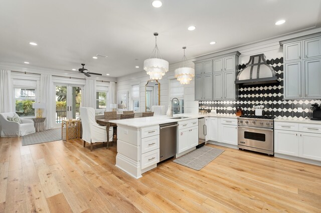 kitchen with custom exhaust hood, hanging light fixtures, light wood-type flooring, ceiling fan with notable chandelier, and appliances with stainless steel finishes