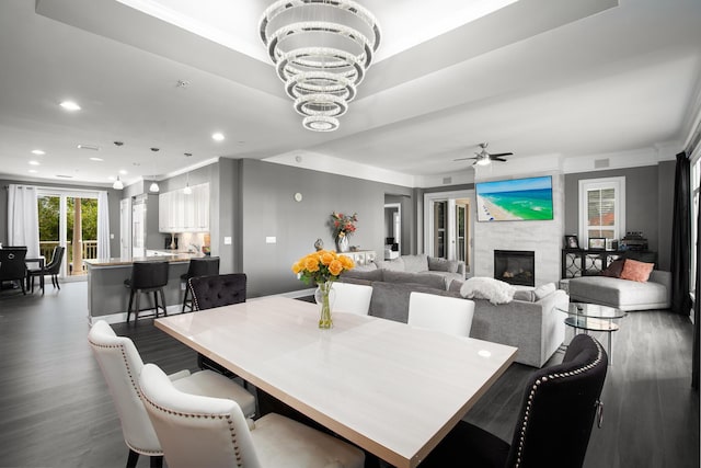 dining space with crown molding, dark hardwood / wood-style flooring, a tiled fireplace, and ceiling fan with notable chandelier