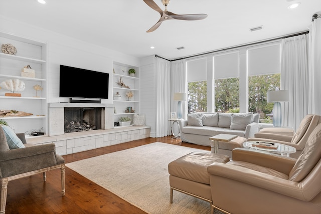 living room with built in features, a tiled fireplace, ceiling fan, and hardwood / wood-style floors