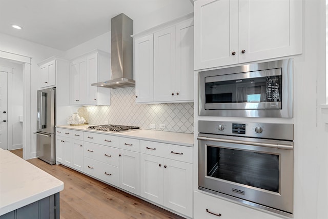 kitchen with wall chimney range hood, light hardwood / wood-style floors, stainless steel appliances, white cabinets, and tasteful backsplash