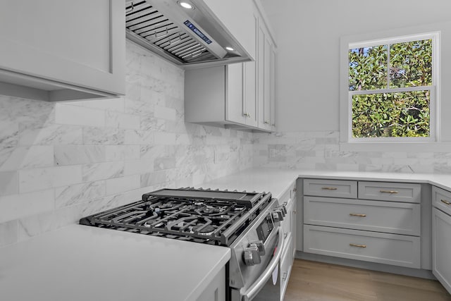 kitchen with tasteful backsplash, wall chimney exhaust hood, stainless steel gas stove, gray cabinets, and light hardwood / wood-style floors