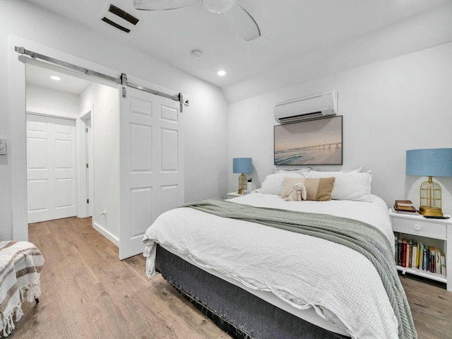 bedroom with a wall mounted AC, wood-type flooring, a barn door, and ceiling fan