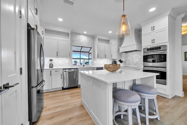 kitchen with appliances with stainless steel finishes, light hardwood / wood-style flooring, a kitchen island, custom range hood, and tasteful backsplash