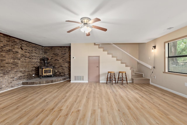 unfurnished living room with ceiling fan, brick wall, a wood stove, and light wood-type flooring