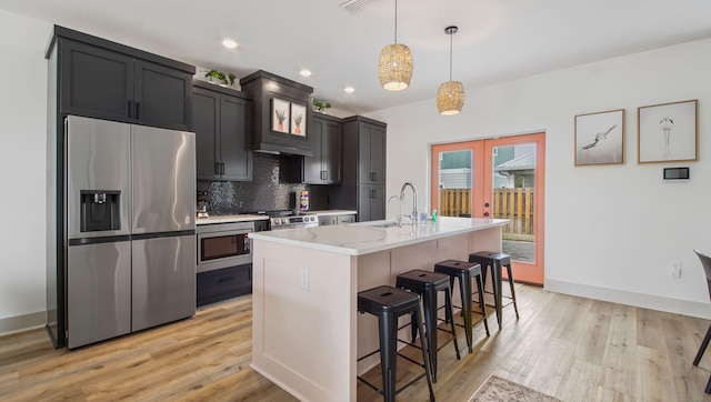 kitchen with appliances with stainless steel finishes, tasteful backsplash, light wood-type flooring, hanging light fixtures, and a kitchen island with sink